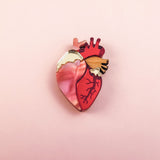 A handcrafted Anatomical Heart Pin, shaped like a human heart and featuring pink, red, and gold tones, rests on a soft pink background—perfect as a unique statement piece.