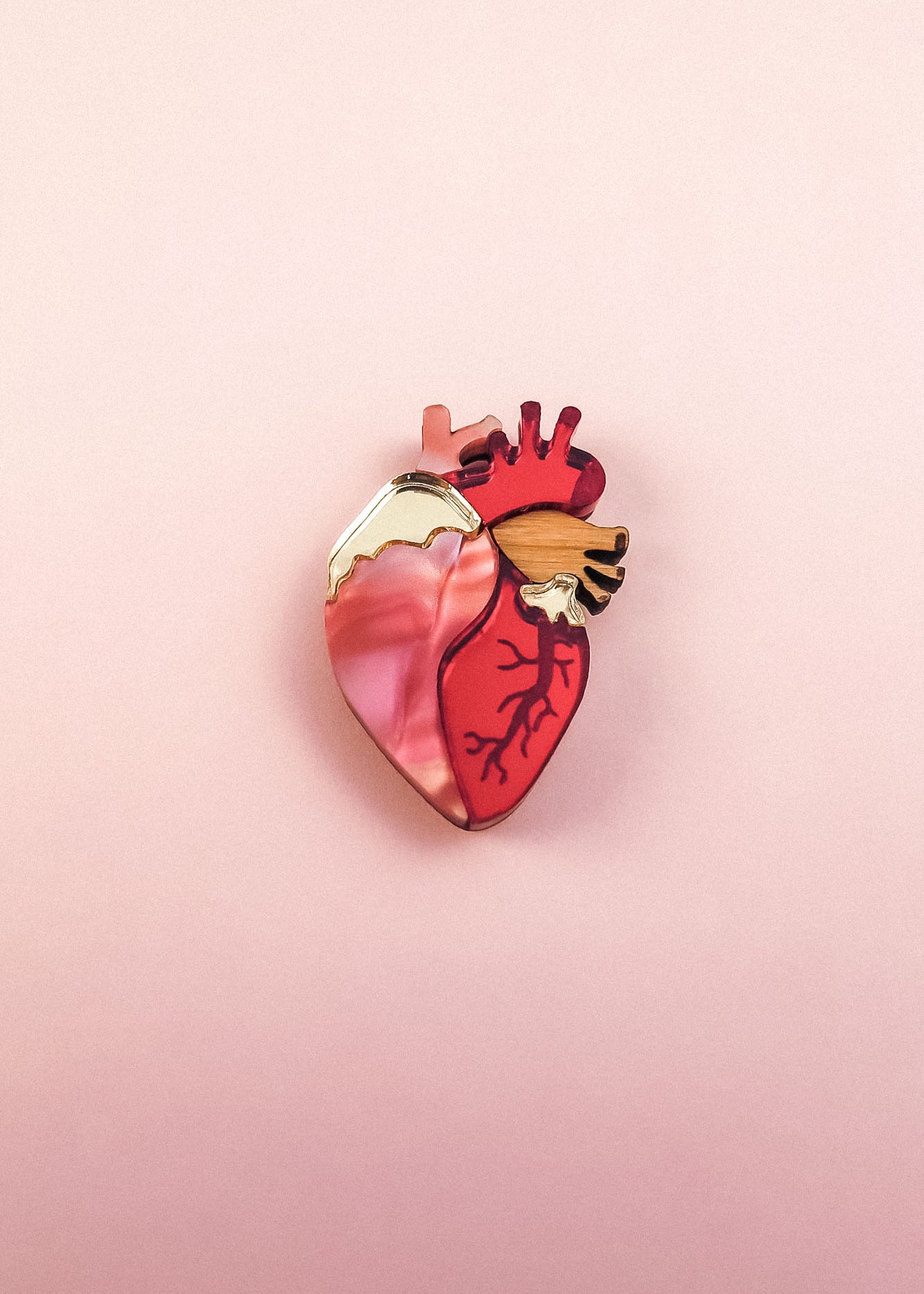 A handcrafted Anatomical Heart Pin, shaped like a human heart and featuring pink, red, and gold tones, rests on a soft pink background—perfect as a unique statement piece.