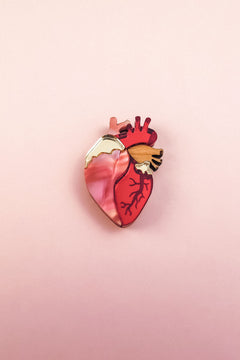 A handcrafted Anatomical Heart Pin, shaped like a human heart and featuring pink, red, and gold tones, rests on a soft pink background—perfect as a unique statement piece.