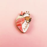 A unique, handcrafted Anatomical Heart Pin with gold accents is displayed on a pink gradient background, making it a striking decorative accessory.
