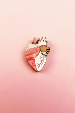 A unique, handcrafted Anatomical Heart Pin with gold accents is displayed on a pink gradient background, making it a striking decorative accessory.
