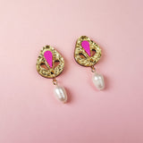 A pair of handcrafted gold-toned drop earrings with pink accents and a dangling white pearl, displayed on a soft pink background.