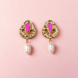 A pair of handcrafted gold drop earrings with intricate designs, featuring a pink enamel detail at the top and a white pearl dangling at the bottom, displayed on a light pink background.
