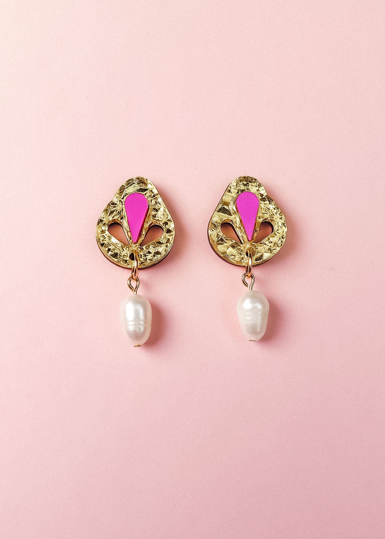A pair of handcrafted gold drop earrings with intricate designs, featuring a pink enamel detail at the top and a white pearl dangling at the bottom, displayed on a light pink background.