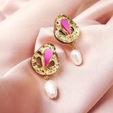 A pair of handcrafted gold drop earrings with intricate designs, pink enamel accents, and white pearl drops, beautifully displayed on soft, pale pink fabric.