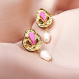 A pair of handcrafted gold drop earrings with pink enamel details and dangling white pearls, displayed on soft pink satin fabric.