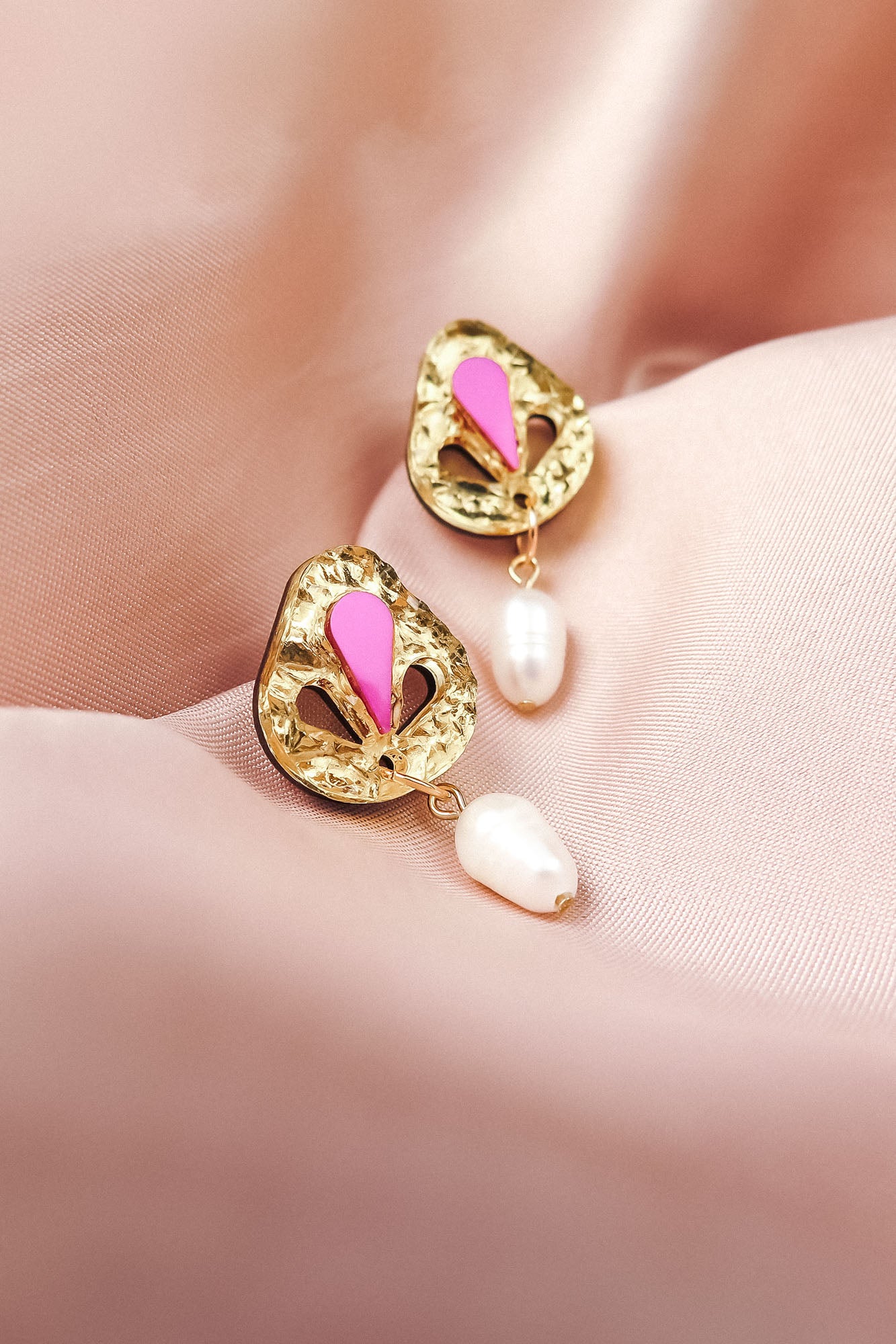 A pair of handcrafted gold drop earrings with pink enamel details and dangling white pearls, displayed on soft pink satin fabric.
