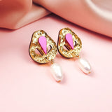 Handcrafted gold drop earrings with textured, abstract shapes, featuring pink and magenta accents and a dangling white pearl—perfect for minimalist fashion—displayed on a soft pink surface with silky pink fabric in the background.
