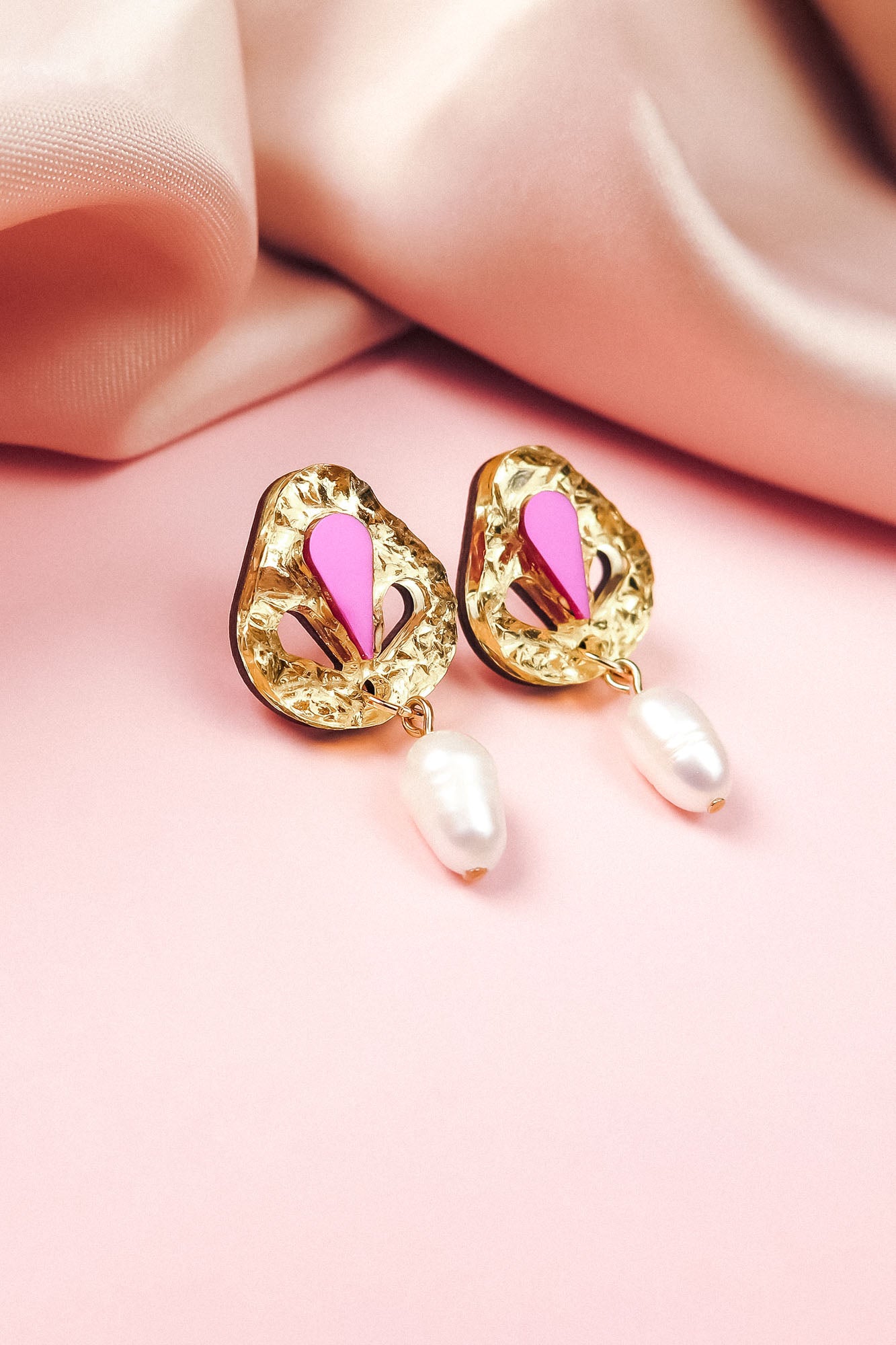 Handcrafted gold drop earrings with textured, abstract shapes, featuring pink and magenta accents and a dangling white pearl—perfect for minimalist fashion—displayed on a soft pink surface with silky pink fabric in the background.
