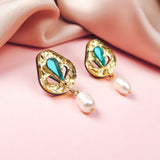 A pair of handcrafted gold earrings with turquoise teardrop accents and dangling white pearls, perfect for minimalist fashion, displayed on a soft pink surface beside a folded blush-colored fabric.