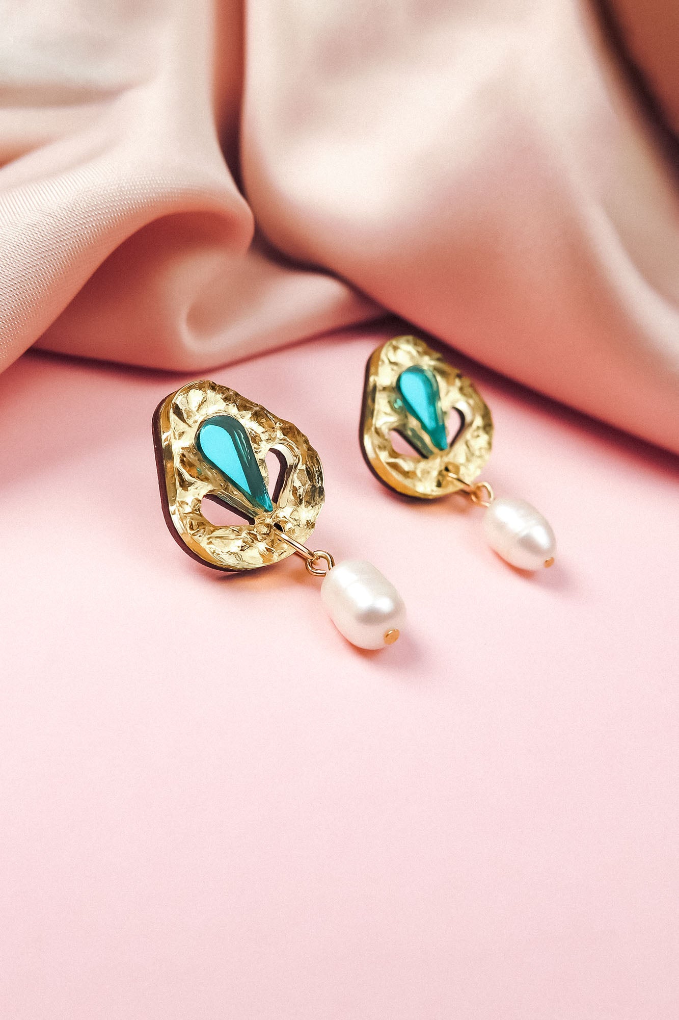 A pair of handcrafted gold earrings with turquoise teardrop accents and dangling white pearls, perfect for minimalist fashion, displayed on a soft pink surface beside a folded blush-colored fabric.