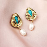 A pair of handcrafted gold earrings with blue enamel accents and dangling white pearls, displayed on light pink satin fabric—perfect for elevating your minimalist fashion style.