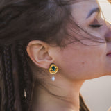 A woman with braided hair poses with her eyes closed, sunlight softly illuminating her face. She wears handcrafted gold earrings featuring a green gem and a dangling pearl, adding an artisanal touch to her minimalist fashion style.