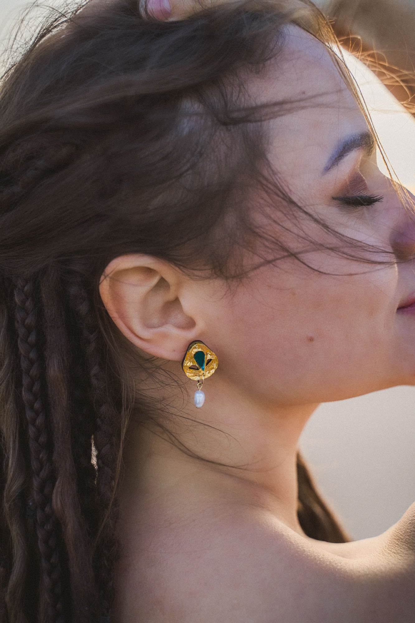 A woman with braided hair poses with her eyes closed, sunlight softly illuminating her face. She wears handcrafted gold earrings featuring a green gem and a dangling pearl, adding an artisanal touch to her minimalist fashion style.