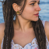 A woman with long, braided hair looks to the side while standing outdoors near water. She wears a floral-patterned dress and Basic Drop Earrings, embracing minimalist fashion against a softly focused background.