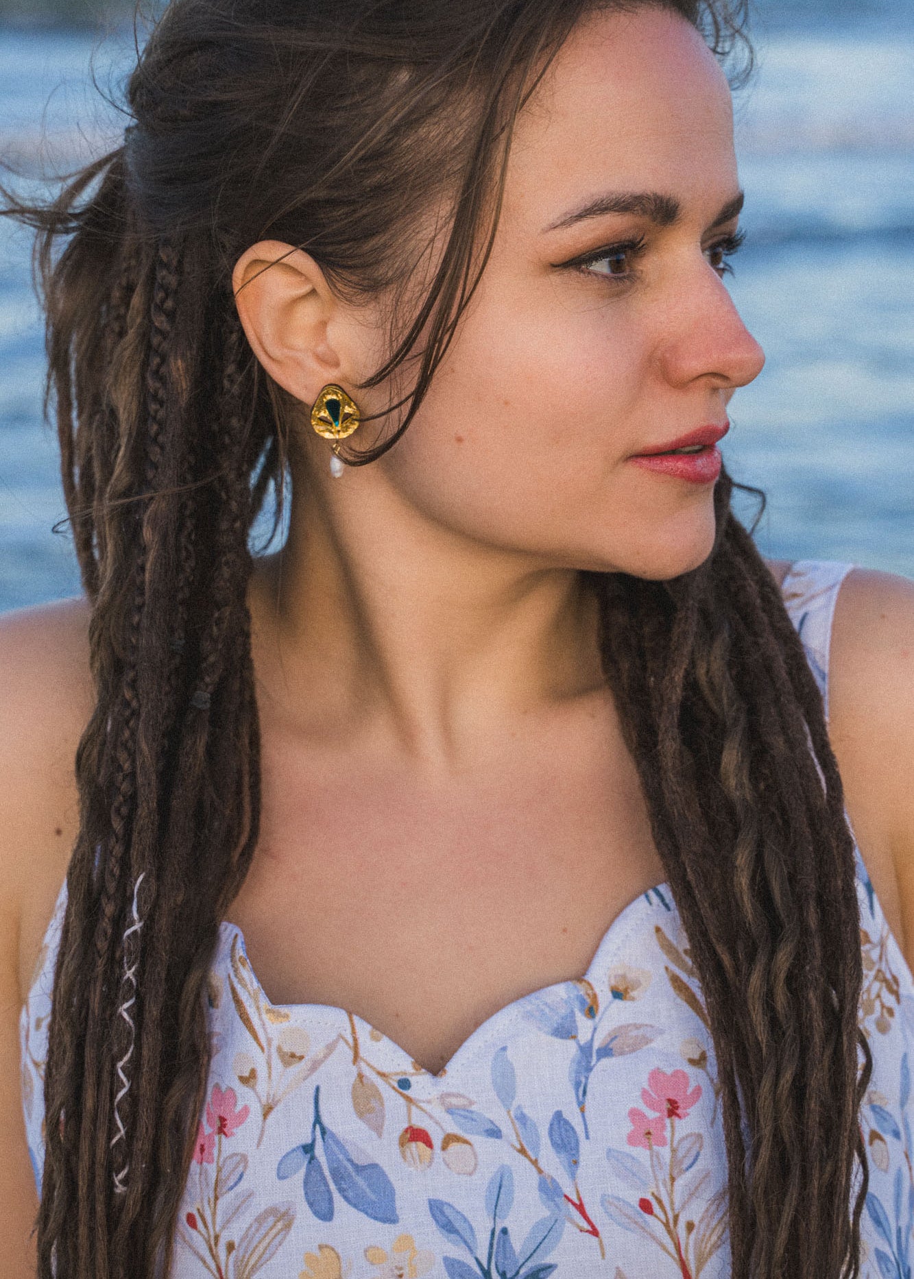A woman with long, braided hair looks to the side while standing outdoors near water. She wears a floral-patterned dress and Basic Drop Earrings, embracing minimalist fashion against a softly focused background.
