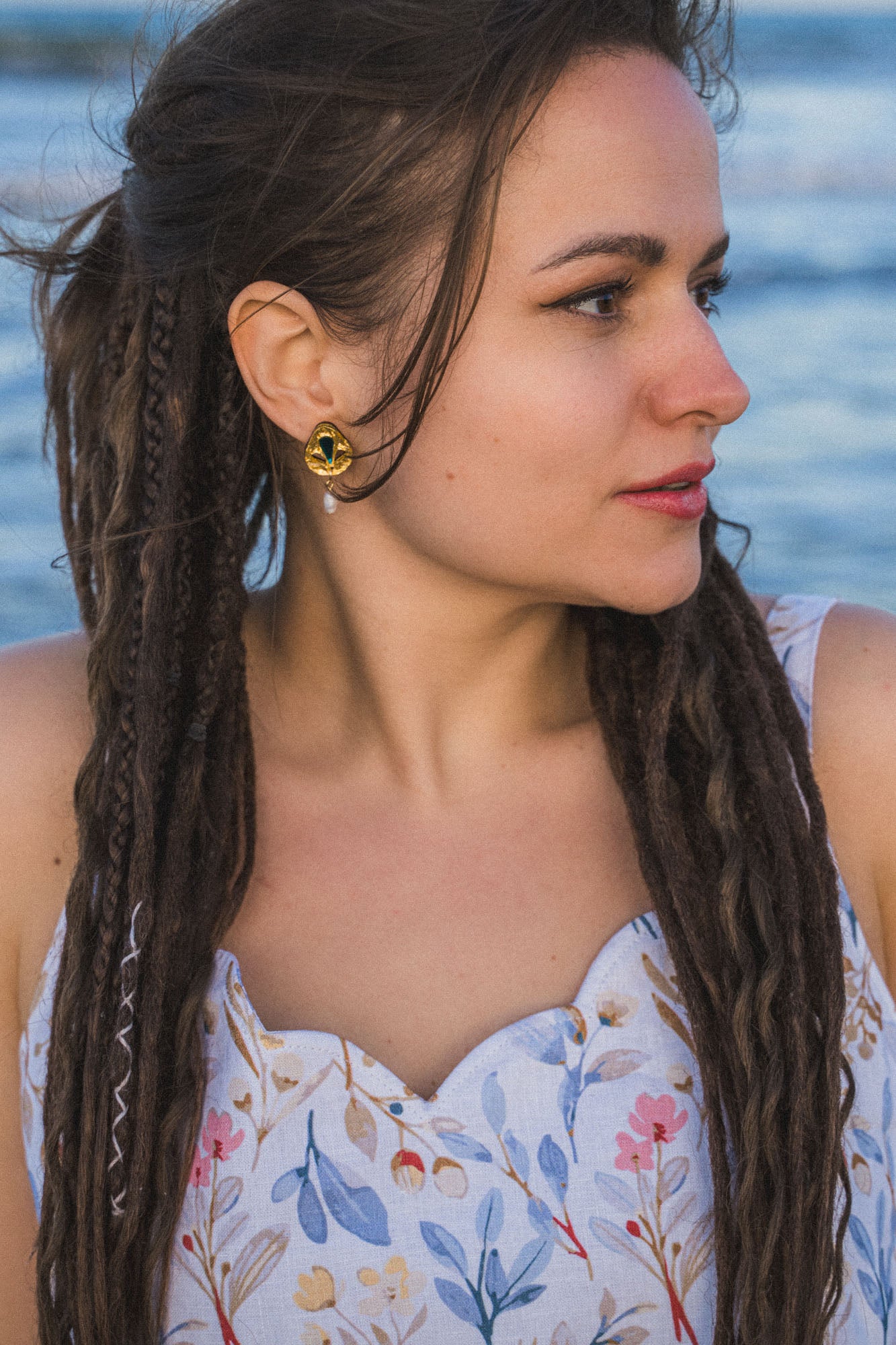 A woman with long, braided hair looks to the side while standing outdoors near water. She wears a floral-patterned dress and Basic Drop Earrings, embracing minimalist fashion against a softly focused background.