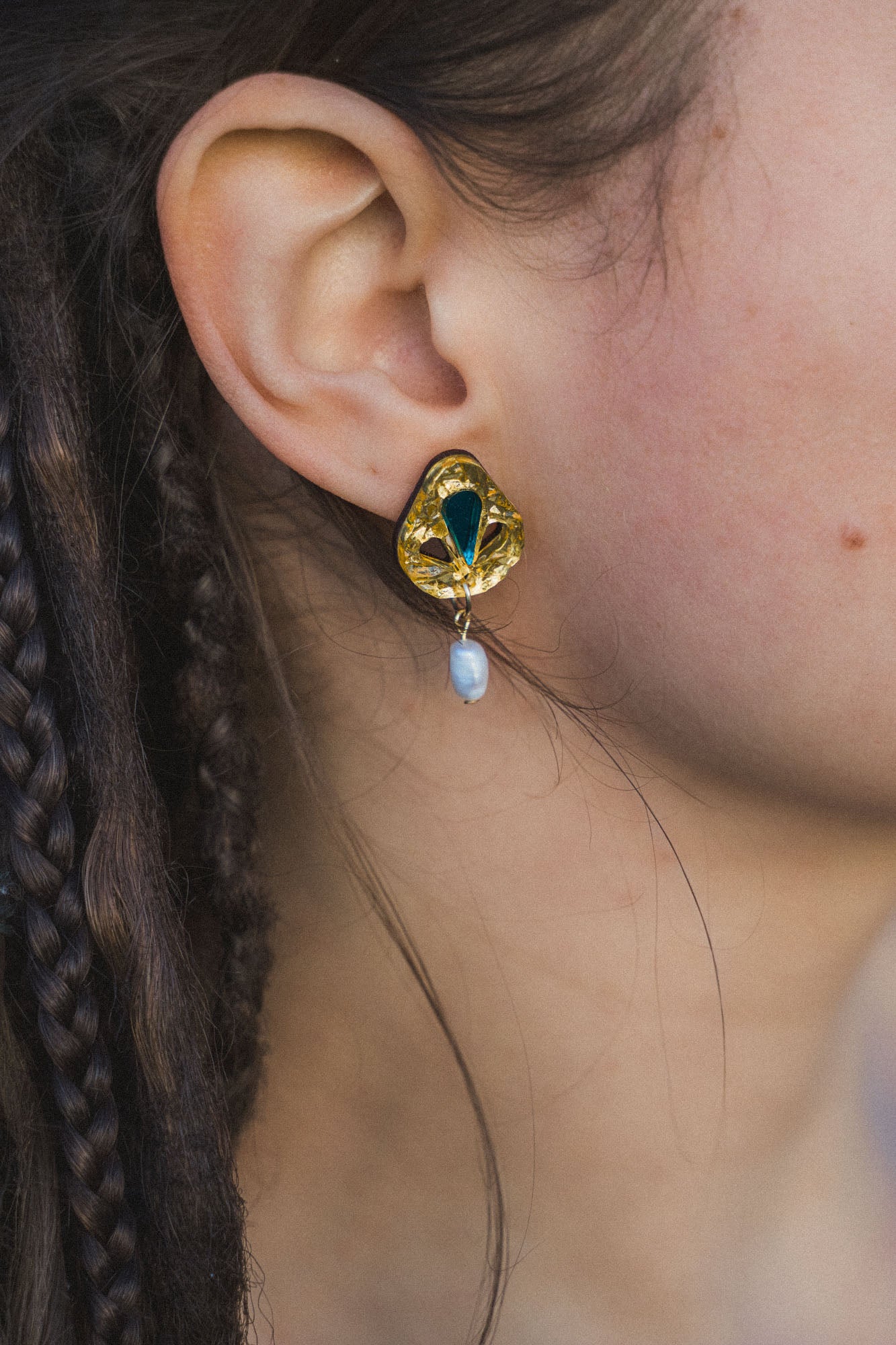 Close-up of a person’s ear wearing handcrafted Basic Drop Earrings featuring a gold setting with a green gemstone and dangling white pearl; the person has braided brown hair, perfect for minimalist fashion lovers.