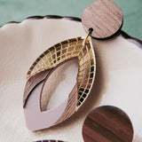 Close-up of handcrafted earrings with circular wooden studs and geometric drop pendants featuring gold lattice, wood accents, and muted purple elements, displayed on a cream-colored ceramic dish. Made with premium acrylic glass details.