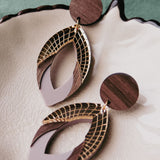 A pair of stylish handcrafted earrings with wooden circular studs and layered geometric dangles featuring gold, brown wood, and light beige accents in premium acrylic glass, displayed on a textured cream-colored dish.