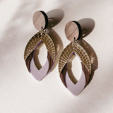 A pair of stylish, handcrafted drop earrings featuring layered geometric shapes in wood, gold, and premium acrylic glass in lavender, casting soft shadows on a light surface.