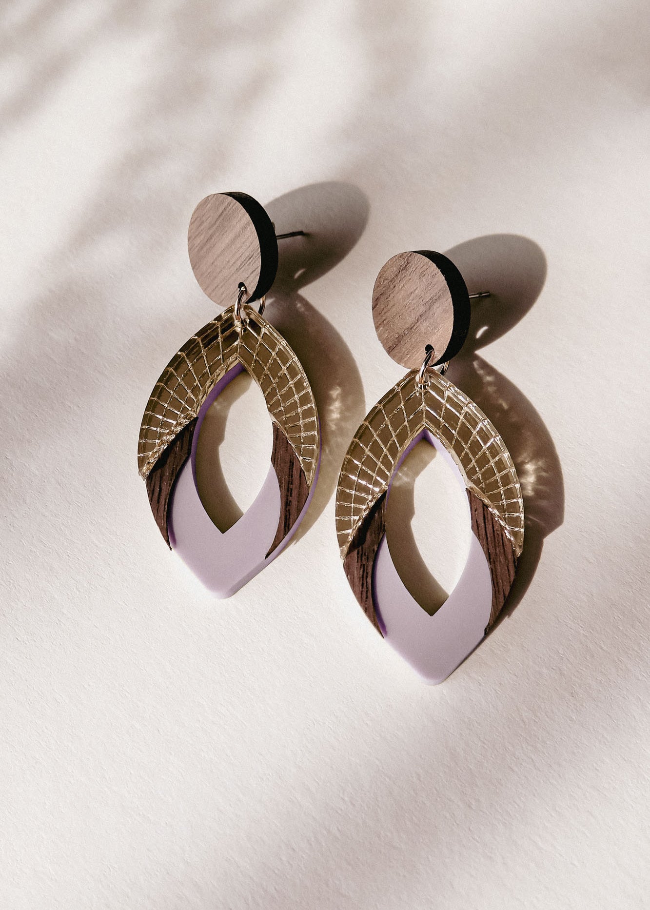 A pair of stylish, handcrafted drop earrings featuring layered geometric shapes in wood, gold, and premium acrylic glass in lavender, casting soft shadows on a light surface.