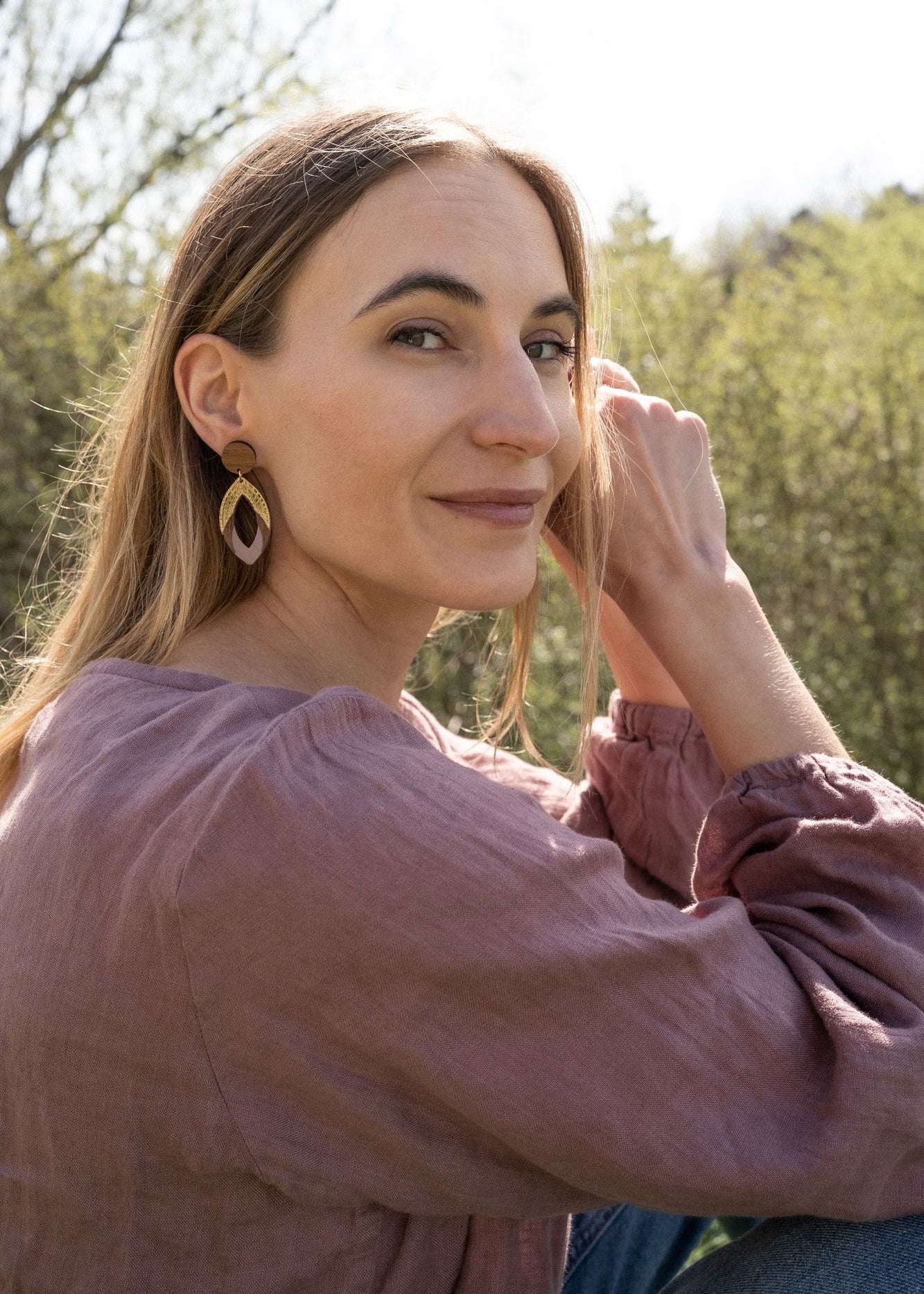 A woman with long light brown hair sits outdoors, smiling slightly as she looks over her shoulder. She wears a mauve blouse and handcrafted Bear Earrings made from premium acrylic glass, with sunlight and greenery in the background.