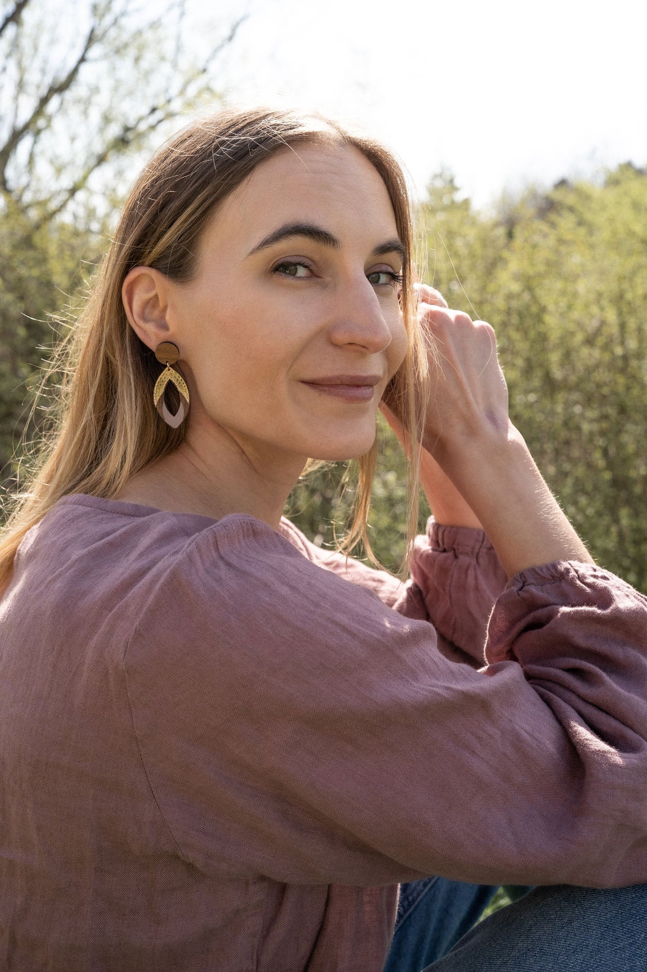 A woman with long light brown hair sits outdoors, smiling slightly as she looks over her shoulder. She wears a mauve blouse and handcrafted Bear Earrings made from premium acrylic glass, with sunlight and greenery in the background.