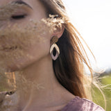 A woman with long hair wearing a mauve top and large, handcrafted bear earrings stands outdoors. Soft sunlight illuminates her face, partially obscured by dried grass in the foreground.