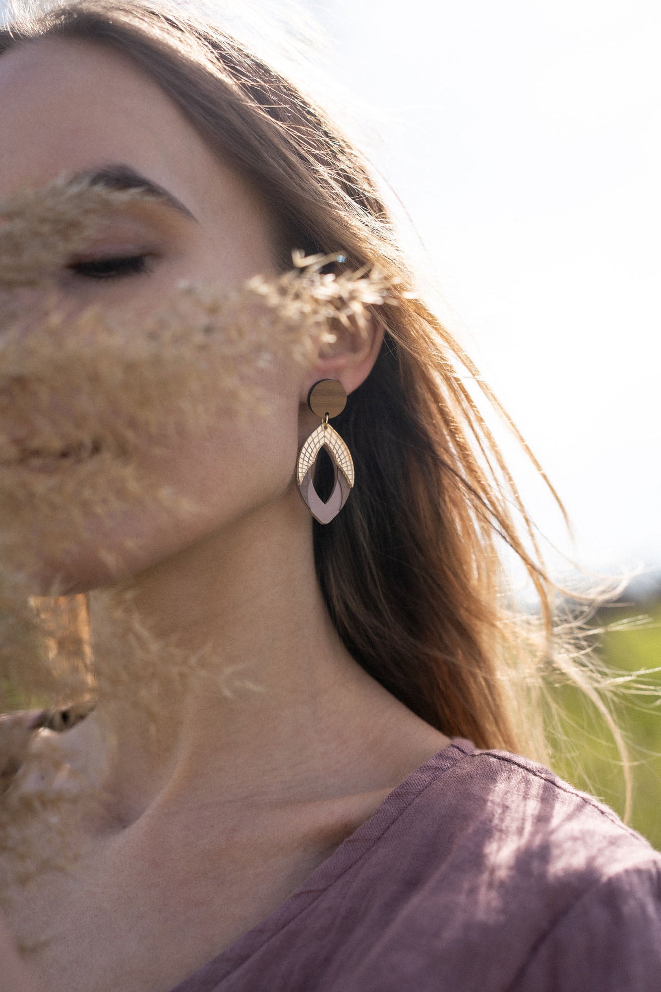A woman with long hair wearing a mauve top and large, handcrafted bear earrings stands outdoors. Soft sunlight illuminates her face, partially obscured by dried grass in the foreground.