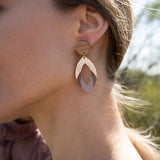 Close-up of a womans ear wearing a large, handcrafted geometric earring featuring wood and gold elements, with a blurred outdoor background.