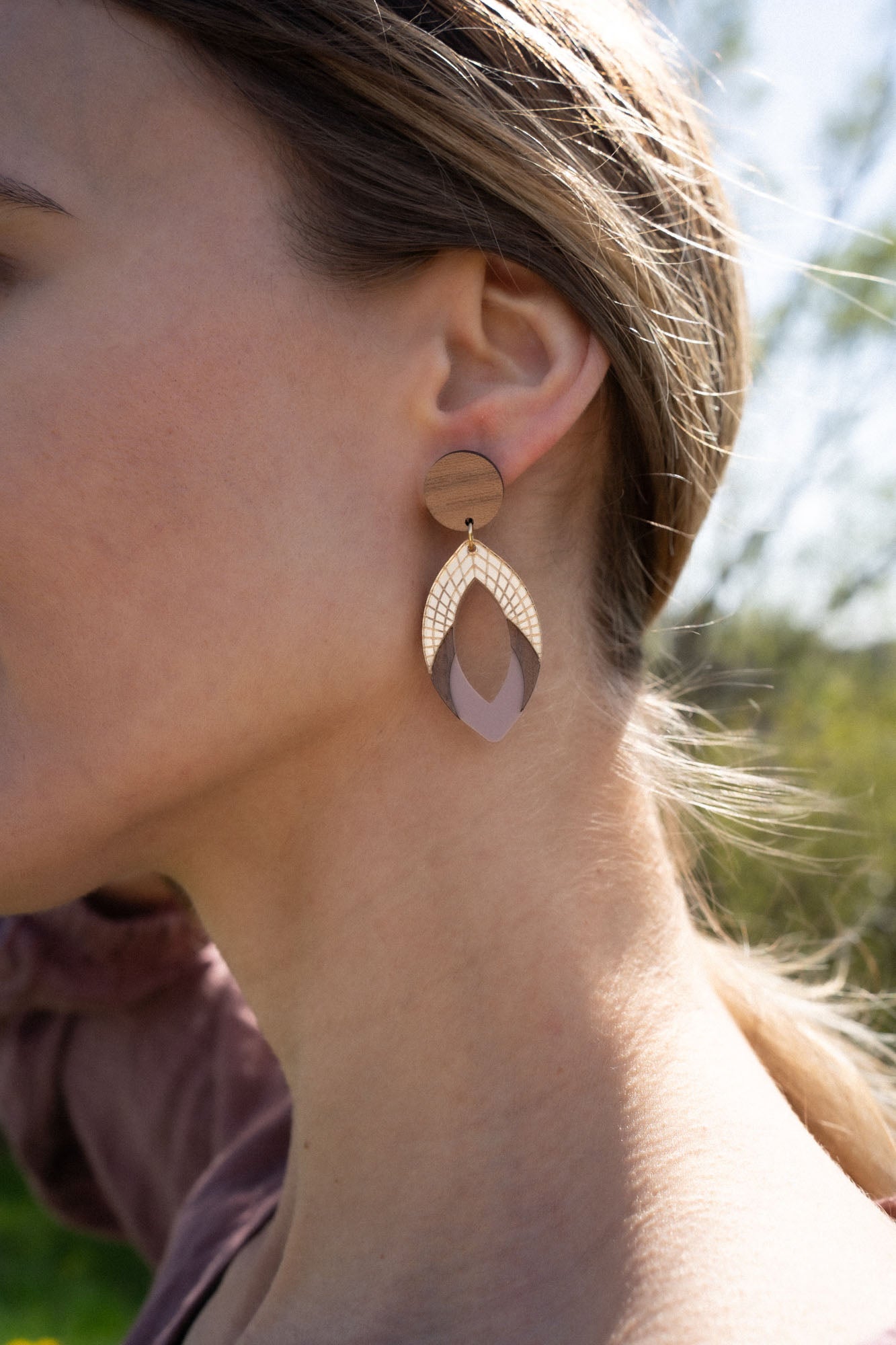 Close-up of a womans ear wearing a large, handcrafted geometric earring featuring wood and gold elements, with a blurred outdoor background.