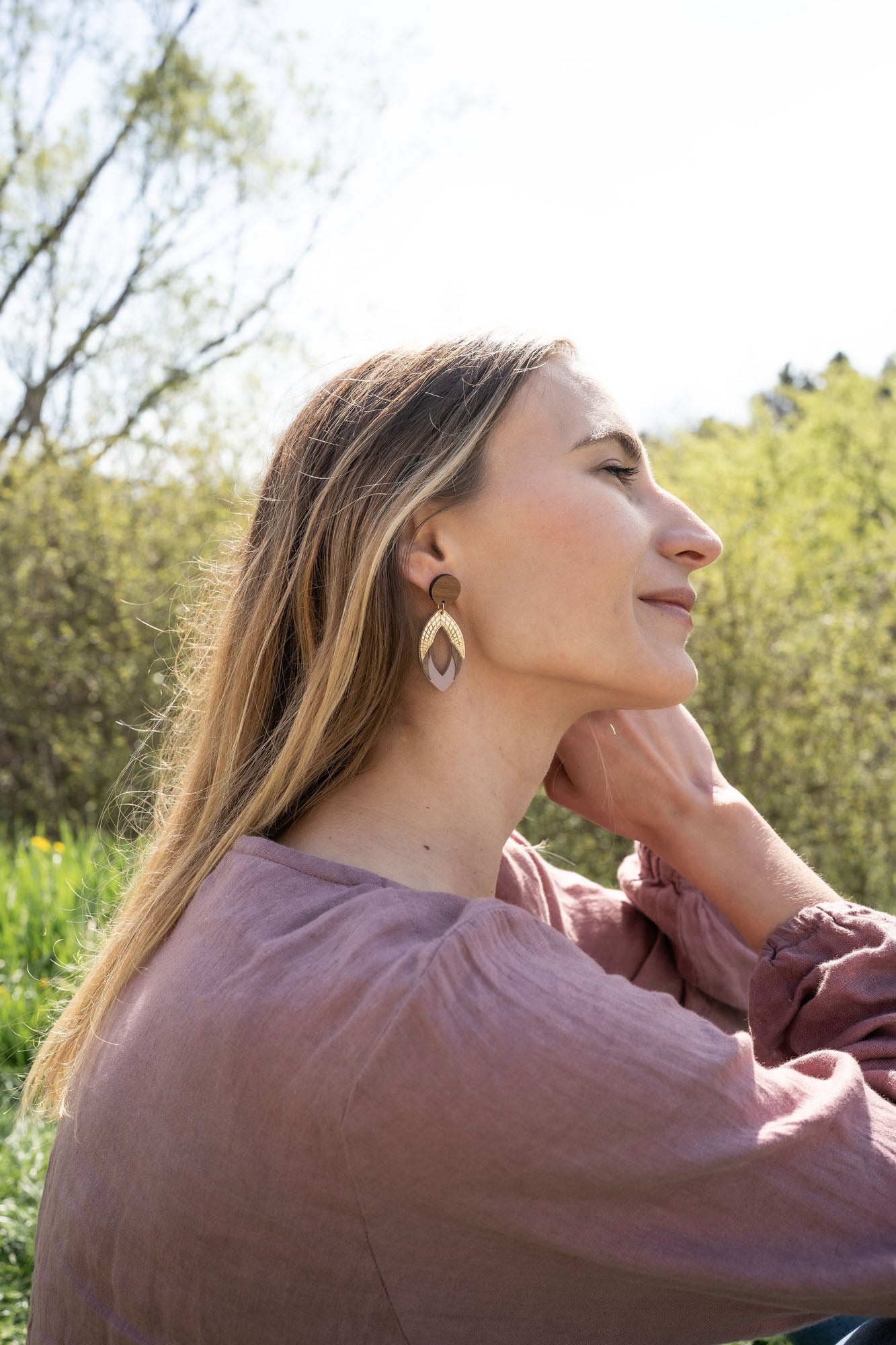 A woman with long hair wearing a mauve top and large handcrafted leaf-shaped earrings sits outdoors, tilting her head back and touching her neck, with greenery and trees blurred in the background.