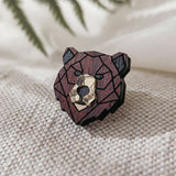 A handcrafted wooden bear brooch in geometric style, featuring dark and light brown tones, rests on textured beige fabric with blurred green fern leaves in the background.
