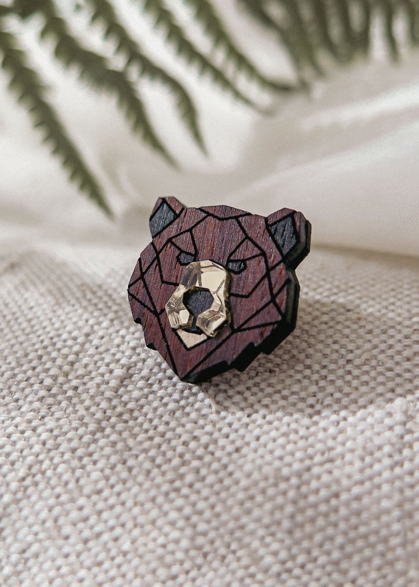 A handcrafted wooden bear brooch in geometric style, featuring dark and light brown tones, rests on textured beige fabric with blurred green fern leaves in the background.
