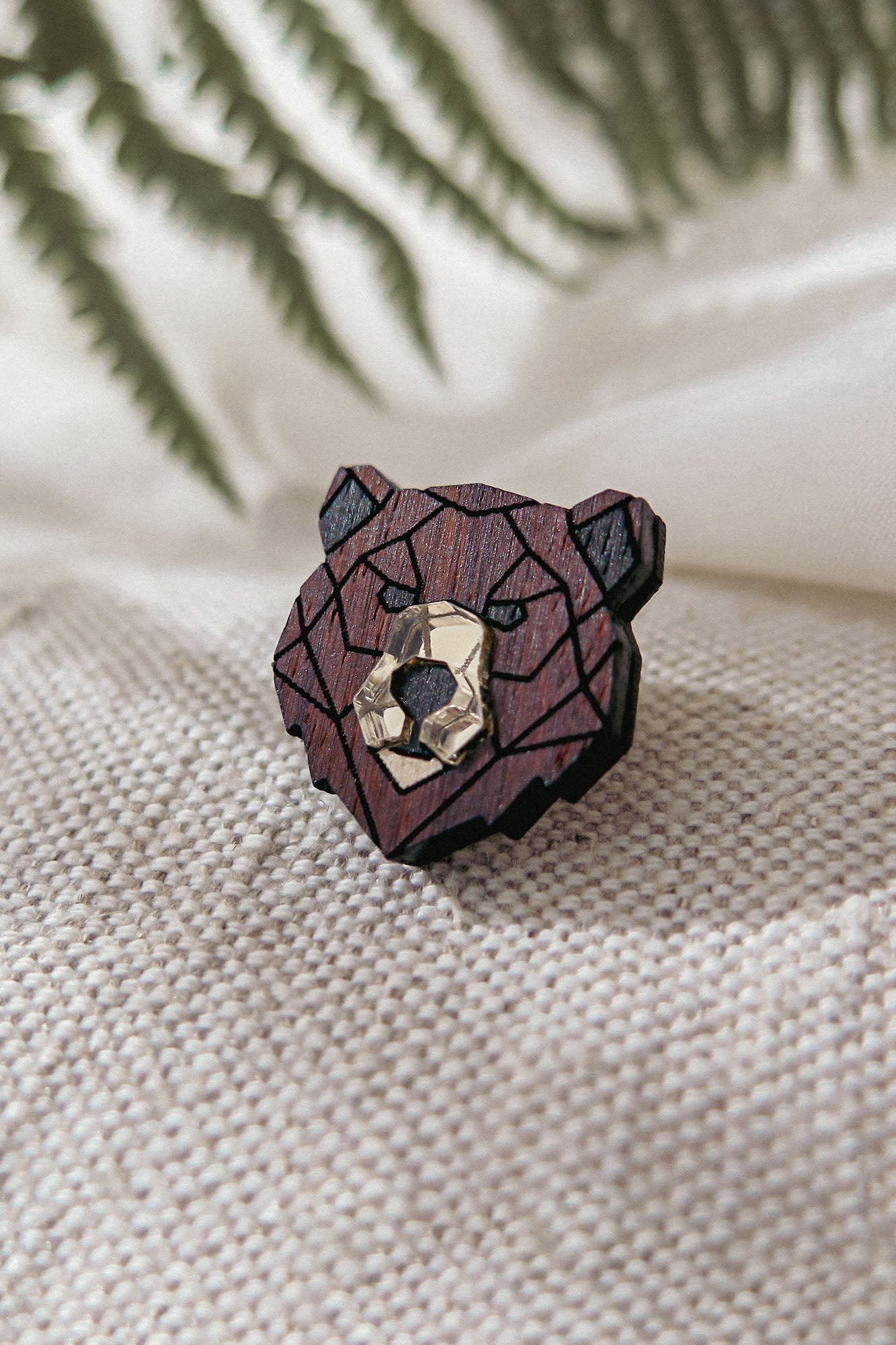 A handcrafted wooden bear brooch in geometric style, featuring dark and light brown tones, rests on textured beige fabric with blurred green fern leaves in the background.