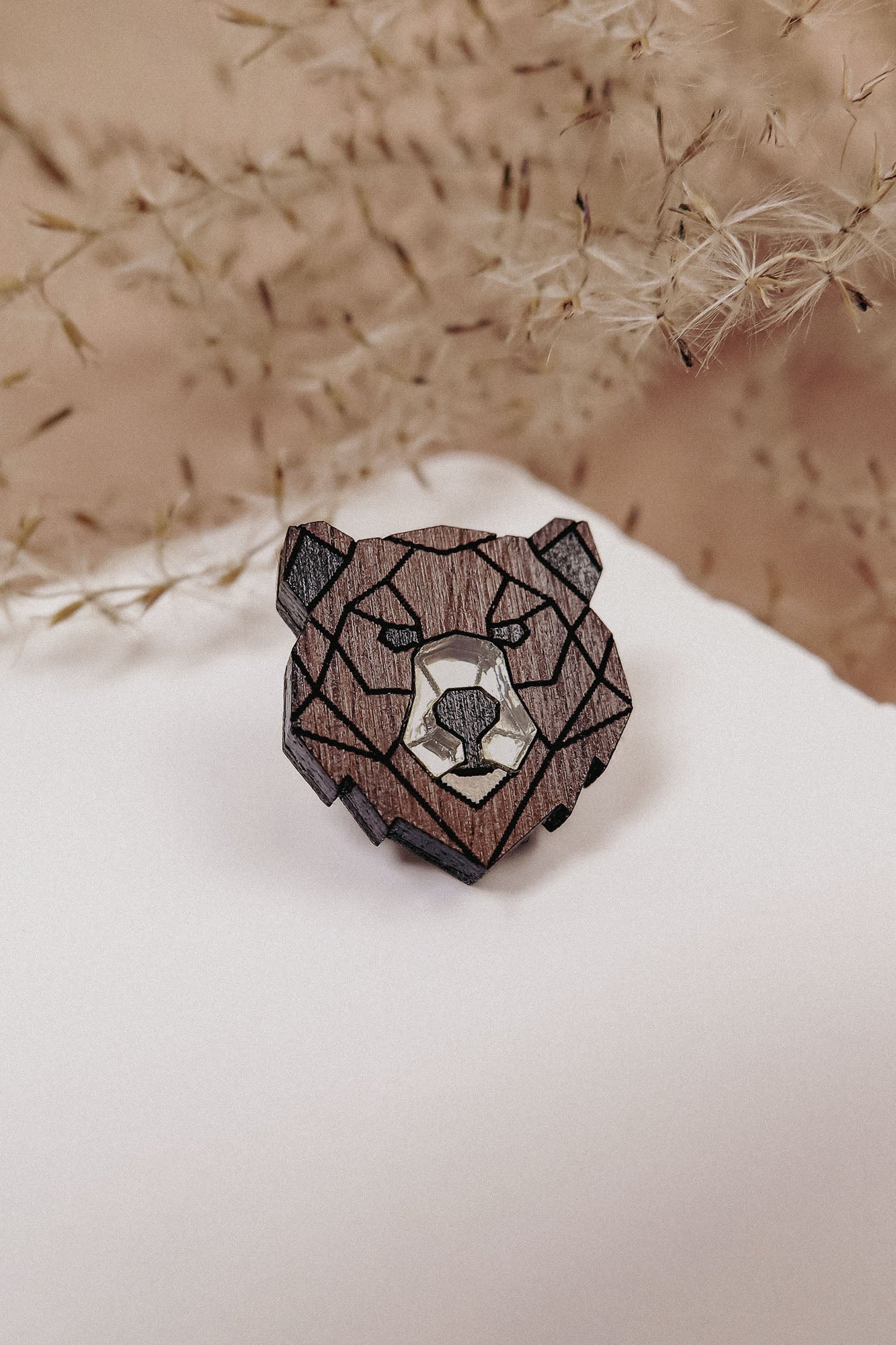 A handcrafted wooden brooch in geometric style, shaped like a bear’s head, rests on a white surface with delicate dried plants softly blurred in the background.