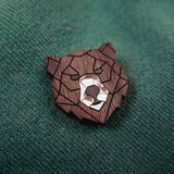 A handcrafted wooden brooch in a geometric style, shaped like a bear’s head, is pinned to green textured fabric. The brooch features angular lines and a white accent on the bear’s snout.