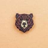 A handcrafted wooden brooch in a geometric style, shaped like a bear’s head with dark brown and light beige sections, placed on a plain light orange background.