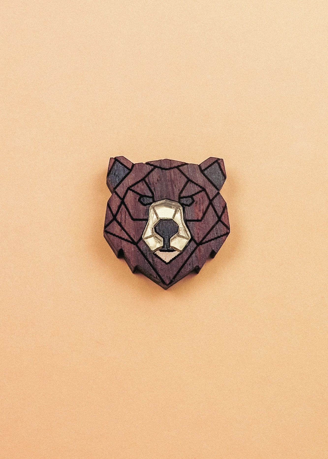 A handcrafted wooden brooch in a geometric style, shaped like a bear’s head with dark brown and light beige sections, placed on a plain light orange background.