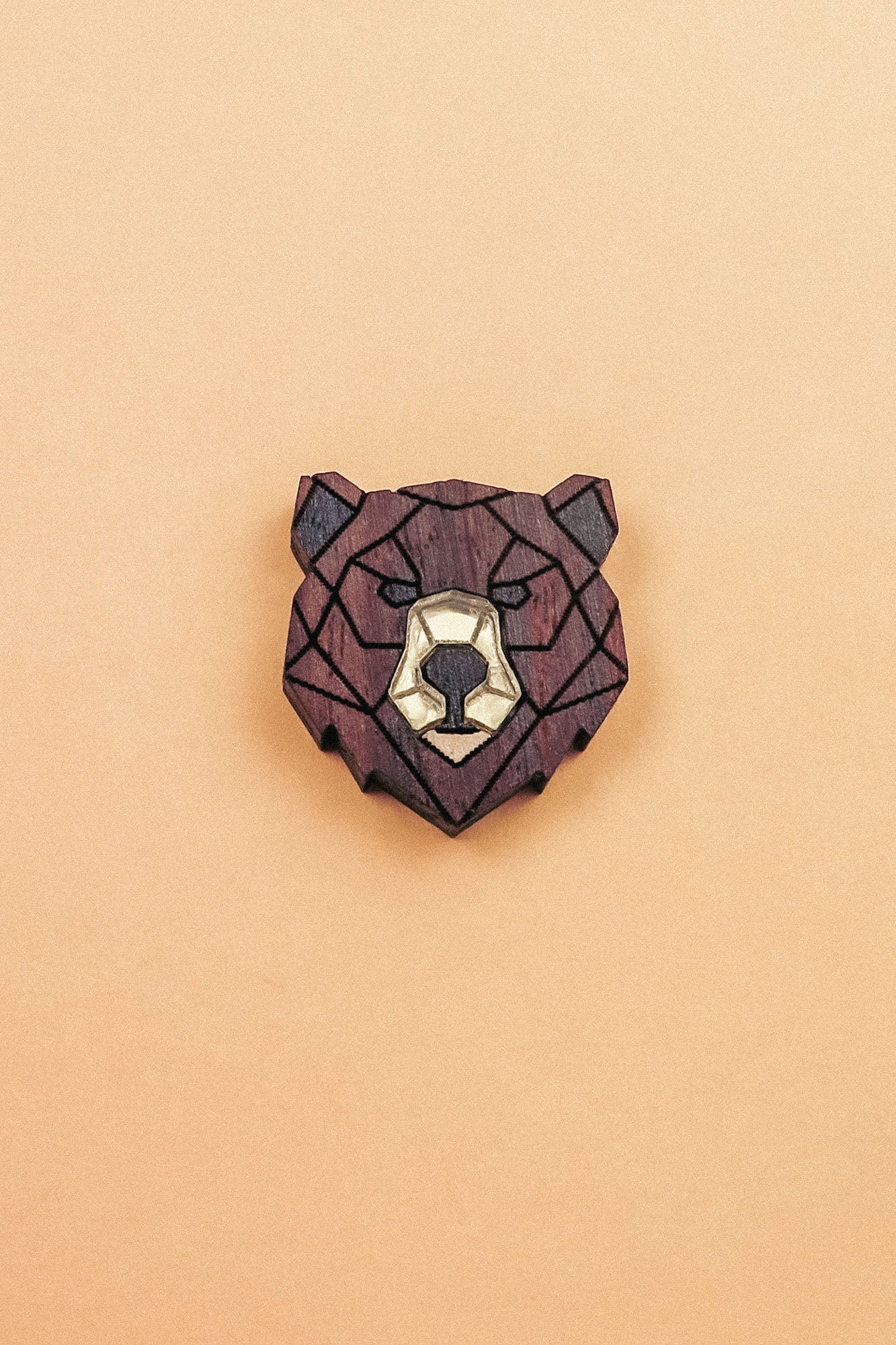 A handcrafted wooden brooch in a geometric style, shaped like a bear’s head with dark brown and light beige sections, placed on a plain light orange background.