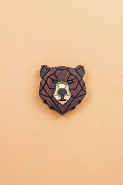 A handcrafted wooden brooch in a geometric style, shaped like a bear’s head with dark brown and light beige sections, placed on a plain light orange background.