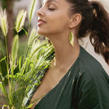 A woman with closed eyes smiles softly while holding green grass stems. She wears a dark green top, unique jewelry with Begonia Leaves Earrings, and stands outdoors in natural light—perfect for plant lovers.