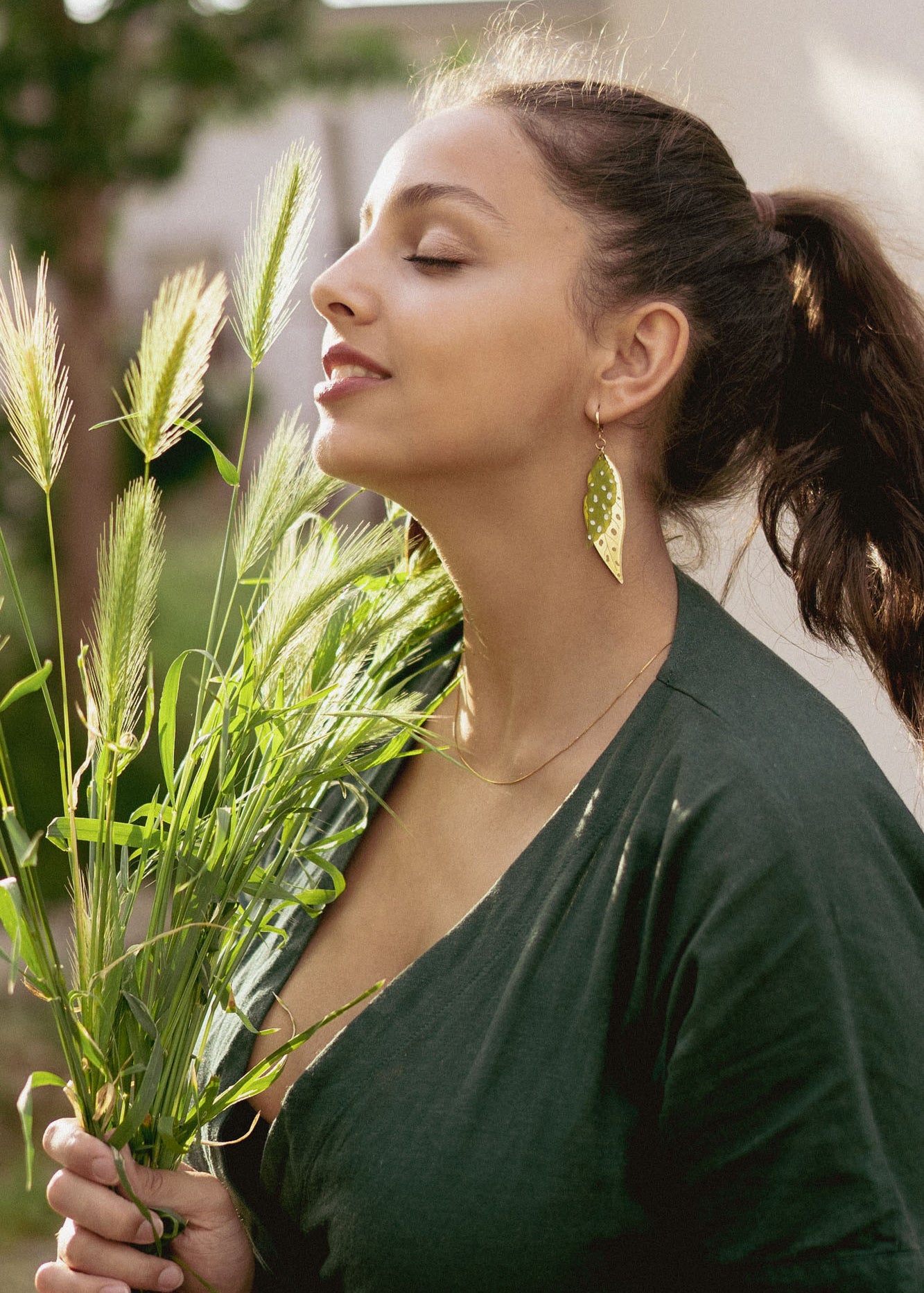 A woman with closed eyes smiles softly while holding green grass stems. She wears a dark green top, unique jewelry with Begonia Leaves Earrings, and stands outdoors in natural light—perfect for plant lovers.