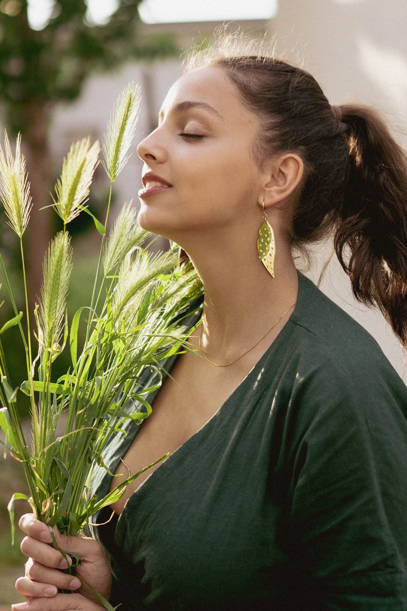 A woman with closed eyes smiles softly while holding green grass stems. She wears a dark green top, unique jewelry with Begonia Leaves Earrings, and stands outdoors in natural light—perfect for plant lovers.