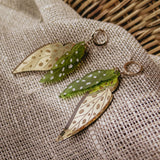A pair of Begonia Leaves Earrings, perfect for plant lovers, shaped like leaves with gold and green hues and white dots, displayed on beige textured fabric next to a woven basket.