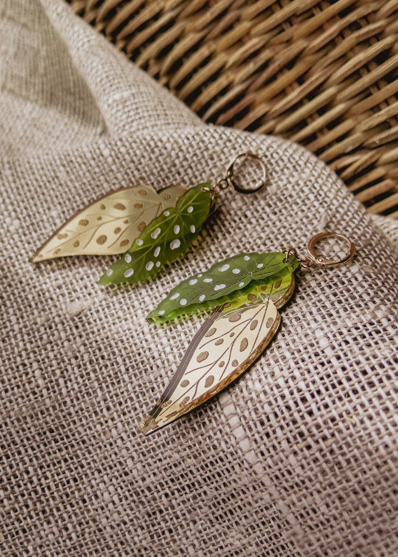 A pair of Begonia Leaves Earrings, perfect for plant lovers, shaped like leaves with gold and green hues and white dots, displayed on beige textured fabric next to a woven basket.