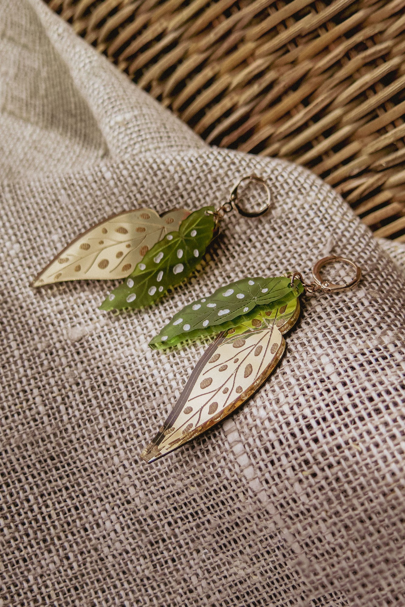 A pair of Begonia Leaves Earrings, perfect for plant lovers, shaped like leaves with gold and green hues and white dots, displayed on beige textured fabric next to a woven basket.