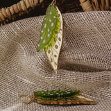 A pair of Begonia Leaves Earrings with green and gold colors and white spots rest on textured beige fabric near a woven basket, making them unique jewelry perfect for plant lovers.
