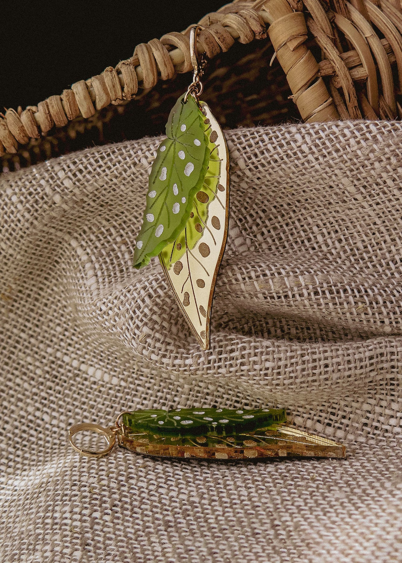 A pair of Begonia Leaves Earrings with green and gold colors and white spots rest on textured beige fabric near a woven basket, making them unique jewelry perfect for plant lovers.